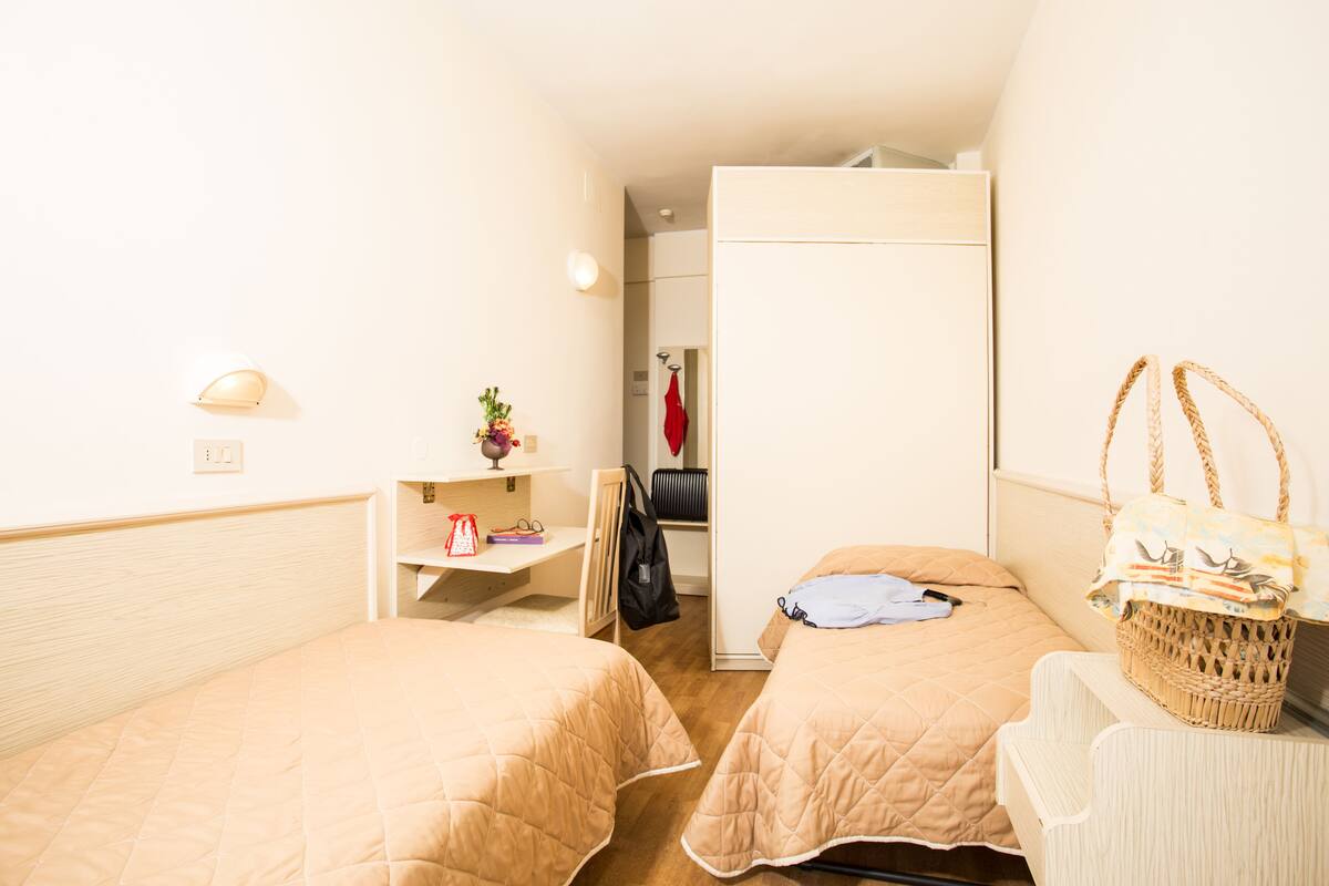 A hotel room with two beds, a desk, a chair, a basket, and a wardrobe.
