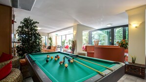 A pool table with balls, a large potted plant, wicker furniture, and a view of the outdoors through large windows. - Hotel Club Misano (Misano Adriatico)