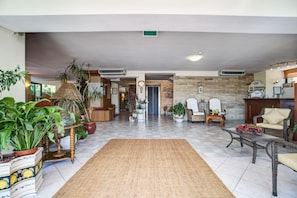 A spacious living area with a tiled floor, potted plants, a large woven rug, and a mix of seating options including a sofa and armchairs. - Hotel Club Misano (Misano Adriatico)