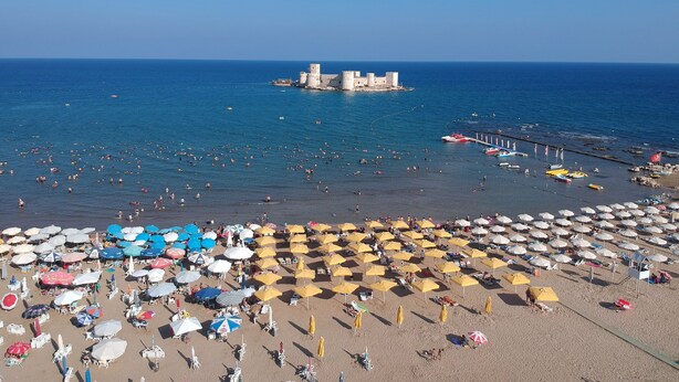 Una playa con numerosos sombrillas y tumbonas, un muelle con barcos y una estructura imponente al estilo de un castillo a lo lejos.