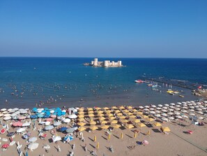A beach with numerous umbrellas and sun loungers, a pier with boats, and a historic castle-like structure in the distance. - Admiral Hotel (Erdemli)