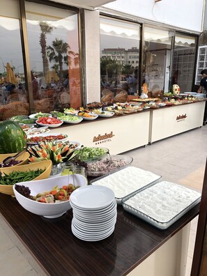 A buffet setup with various food items and a watermelon centerpiece, located in a hotel with a view of the cityscape through large windows. - Admiral Hotel (Erdemli)