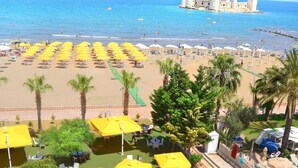 Un área de piscina con sombrillas amarillas y sillas de descanso, una playa con sombrillas y gente, y una estructura que se asemeja a un castillo a lo lejos.
