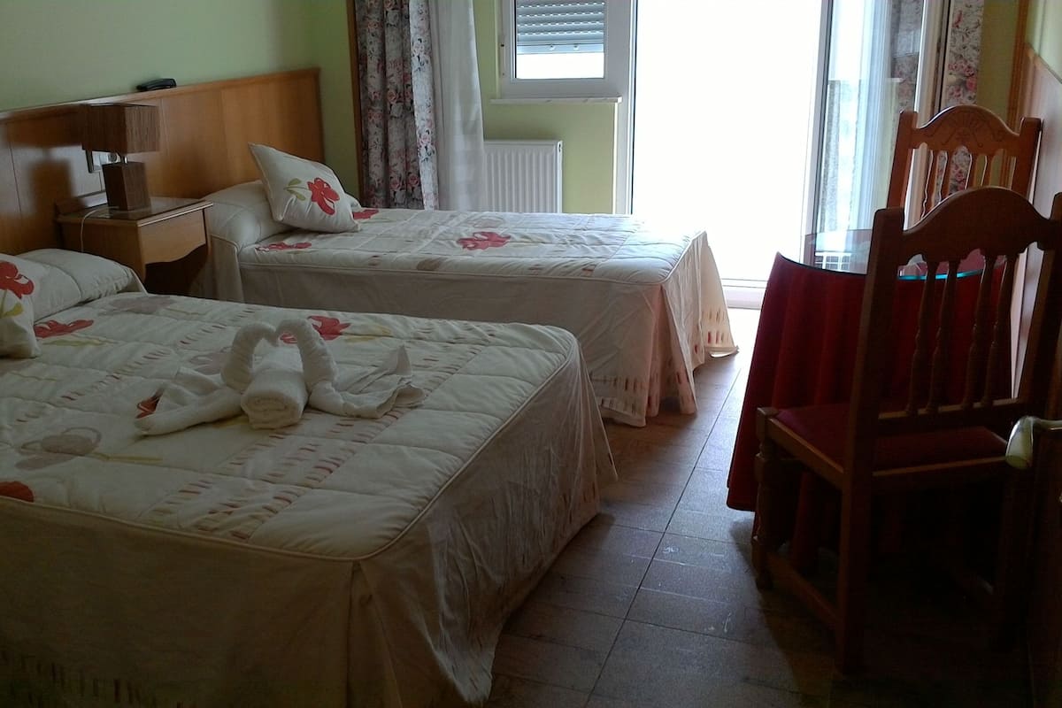 Dos camas con ropa de cama y toallas estampadas, una mesita de noche de madera, una silla y una ventana con cortinas.