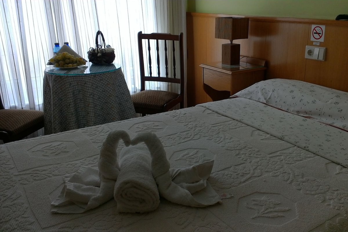 Una habitación de hotel con una cama, una silla, una mesa con una cesta y botellas, y una mesita de noche con una lámpara.