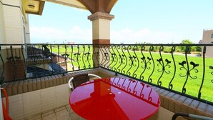 Un balcón con una mesa y sillas rojas, vista a un césped verde y un edificio al fondo.