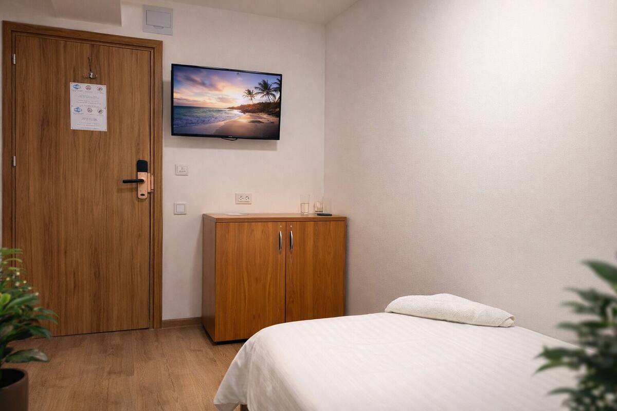 Un dormitorio con un armario de madera, una cama con edredón blanco, un televisor en la pared mostrando una escena de playa, y un mueble pequeño con vasos encima.