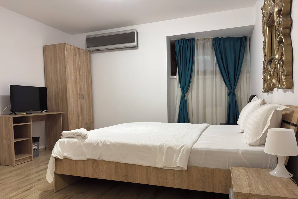 Un dormitorio moderno con cama de madera, un televisor sobre un mueble de madera, un armario y una ventana con cortinas azules.