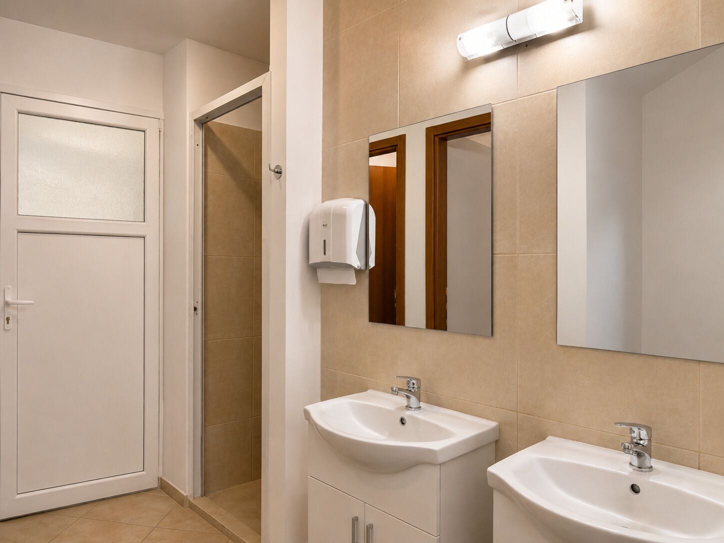 A bathroom with two sinks, a mirror, a towel dispenser, and a door.