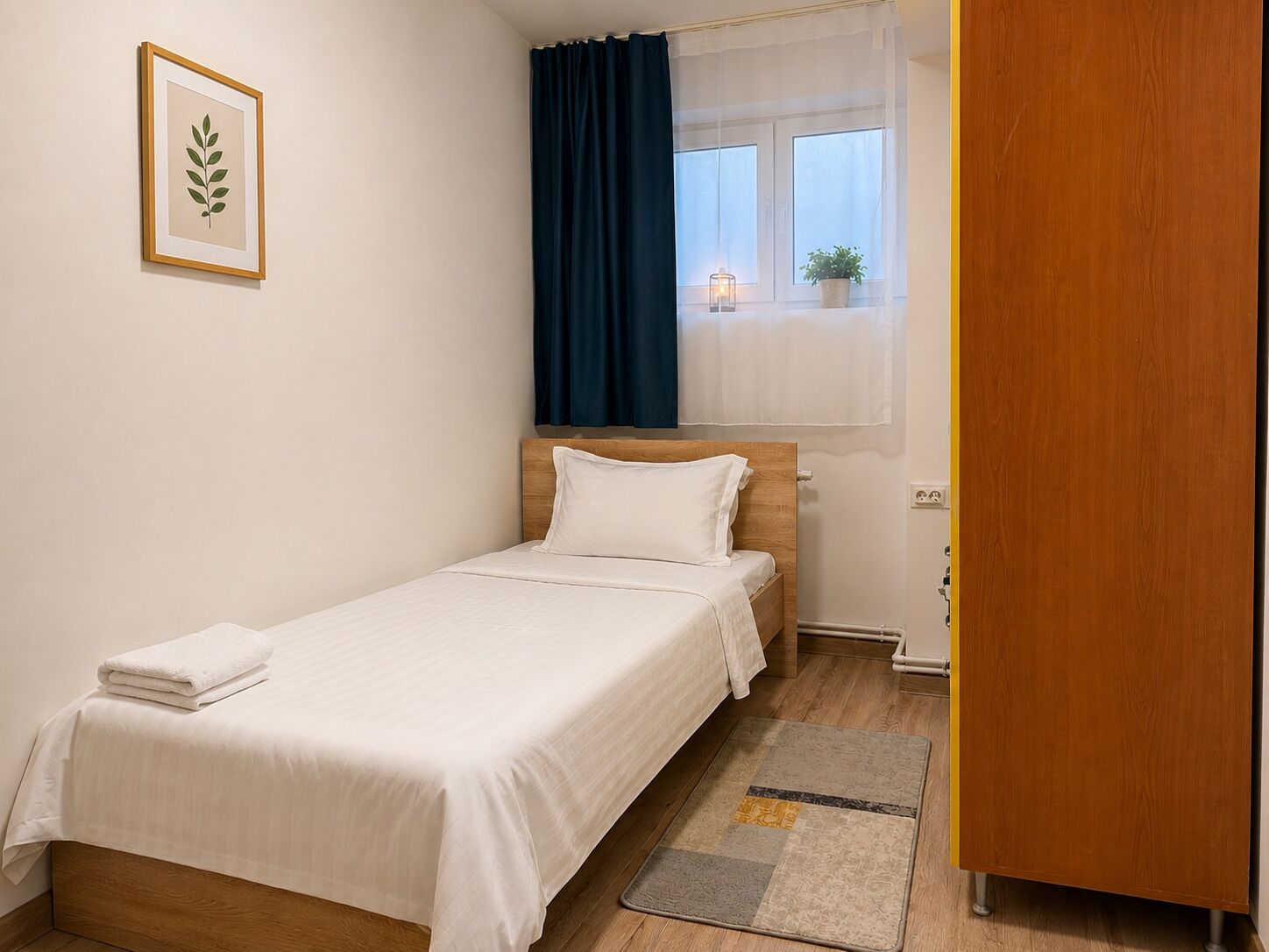 A single bed with white bedding, a bedside table, a nightstand, a window with curtains, a framed picture on the wall, and a wooden wardrobe.