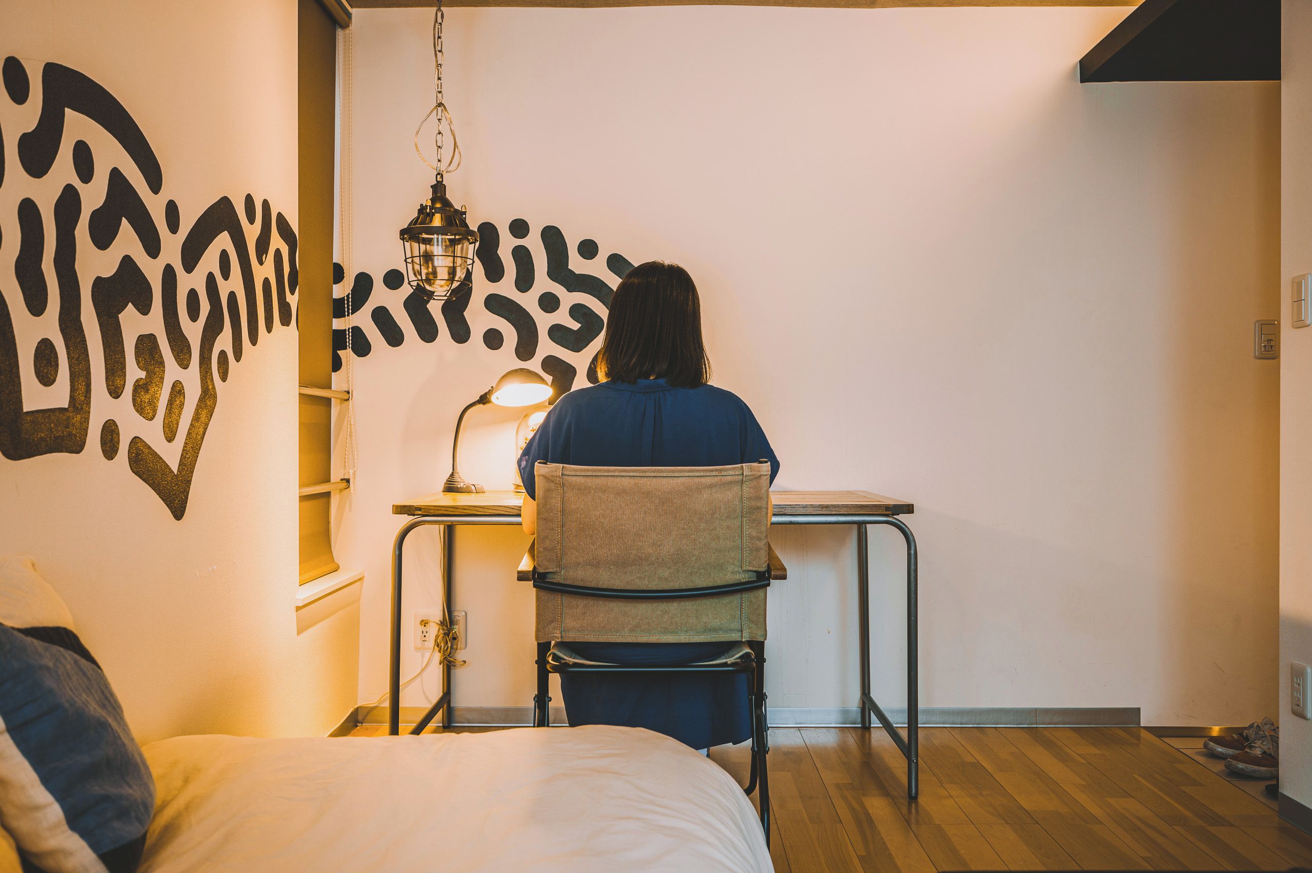 A person seated at a desk in a room with a bed, a wall with abstract art, and a hanging light fixture.