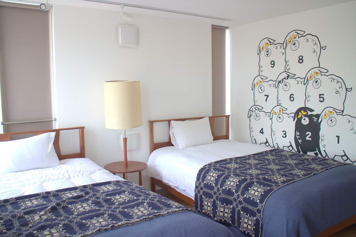 Une chambre d’hôtel avec deux lits, une table de chevet avec une lampe et une fresque murale représentant des moutons avec des chiffres.