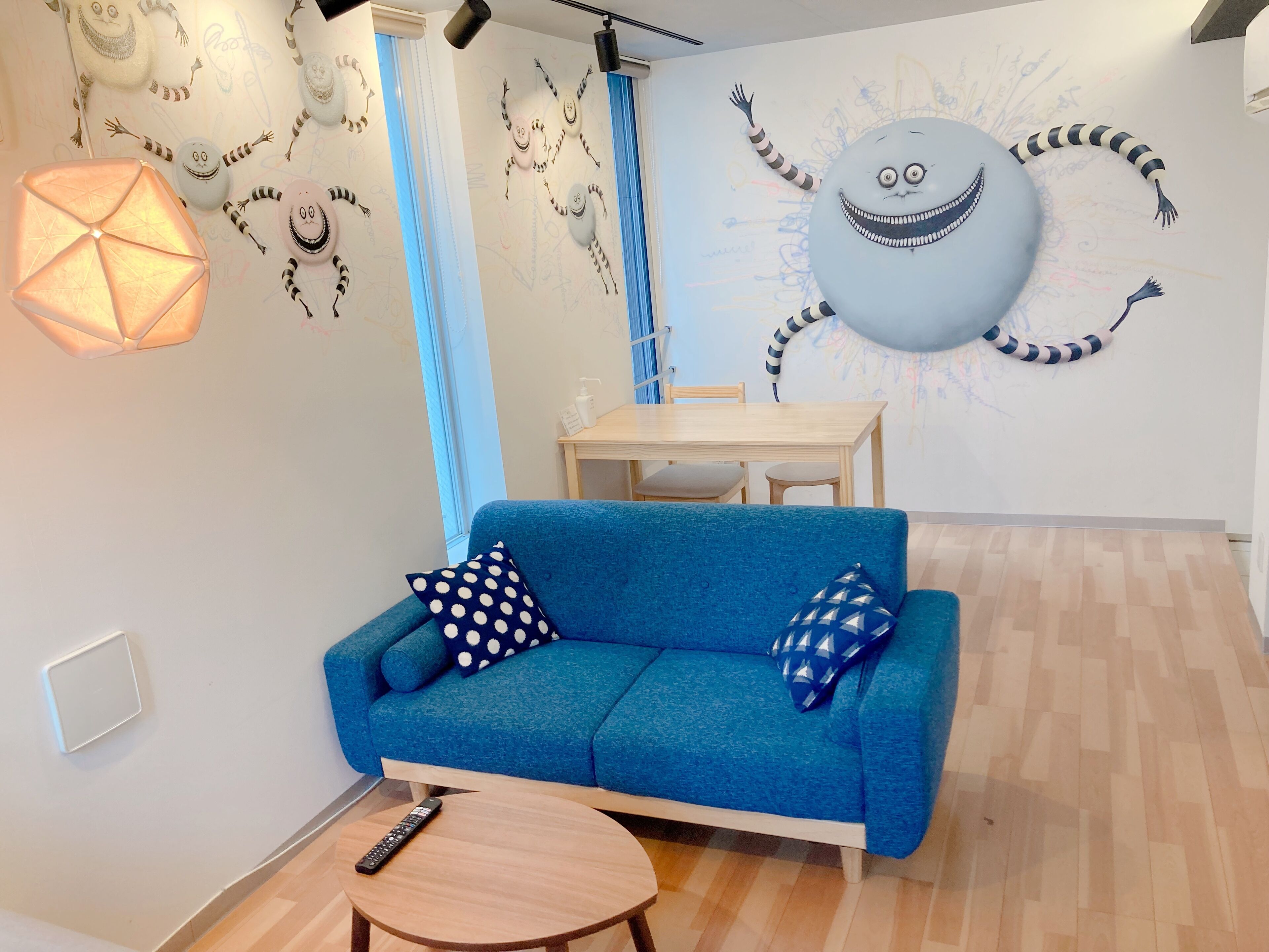 Habitación con un sofá azul, dos almohadones estampados, una pequeña mesa de madera, un control remoto y una pared con un gran mural de un personaje de caricatura.