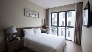 A hotel room with a bed, a nightstand with a lamp, a phone, a framed picture on the wall, a window with curtains, and a TV mounted on the wall. - Tigon Hotel (Hanoi)