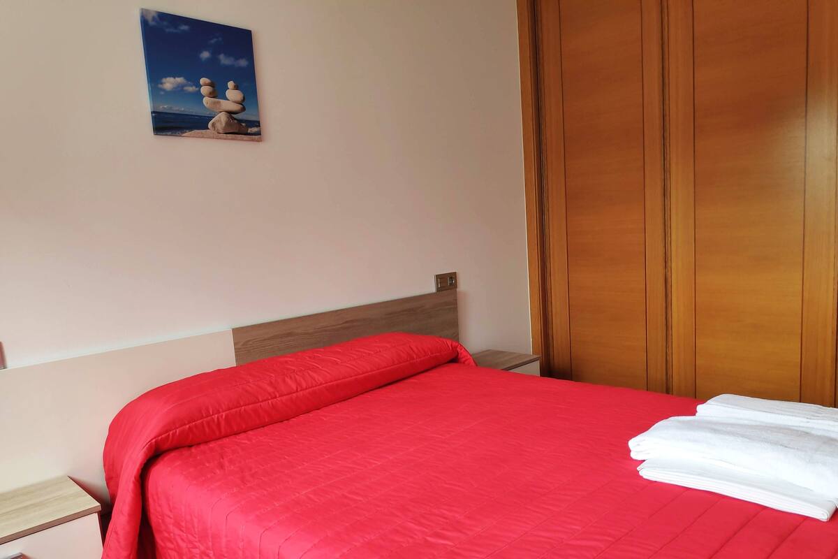 A hotel room with a red bedspread, wooden headboard, a wooden wardrobe, and a small painting on the wall.