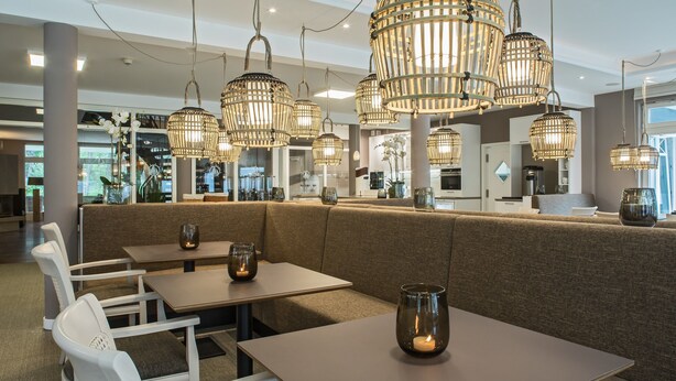 A modern restaurant interior with high ceilings, wicker pendant lighting, cushioned seating, and glass candle holders on tables. - Upstalsboom Trassenheide (Trassenheide)