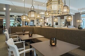 A modern restaurant interior with high ceilings, wicker pendant lighting, cushioned seating, and glass candle holders on tables. - Upstalsboom Trassenheide (Trassenheide)