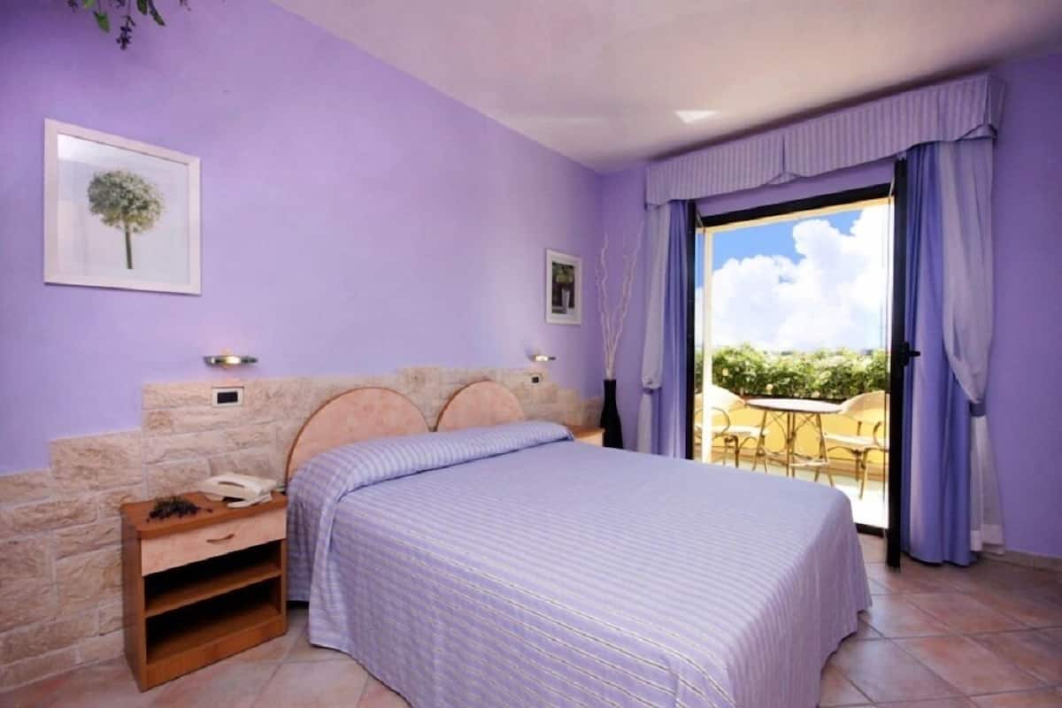 A bedroom with a bed, a nightstand, a stone wall, a window with curtains, and a view of the outdoors.