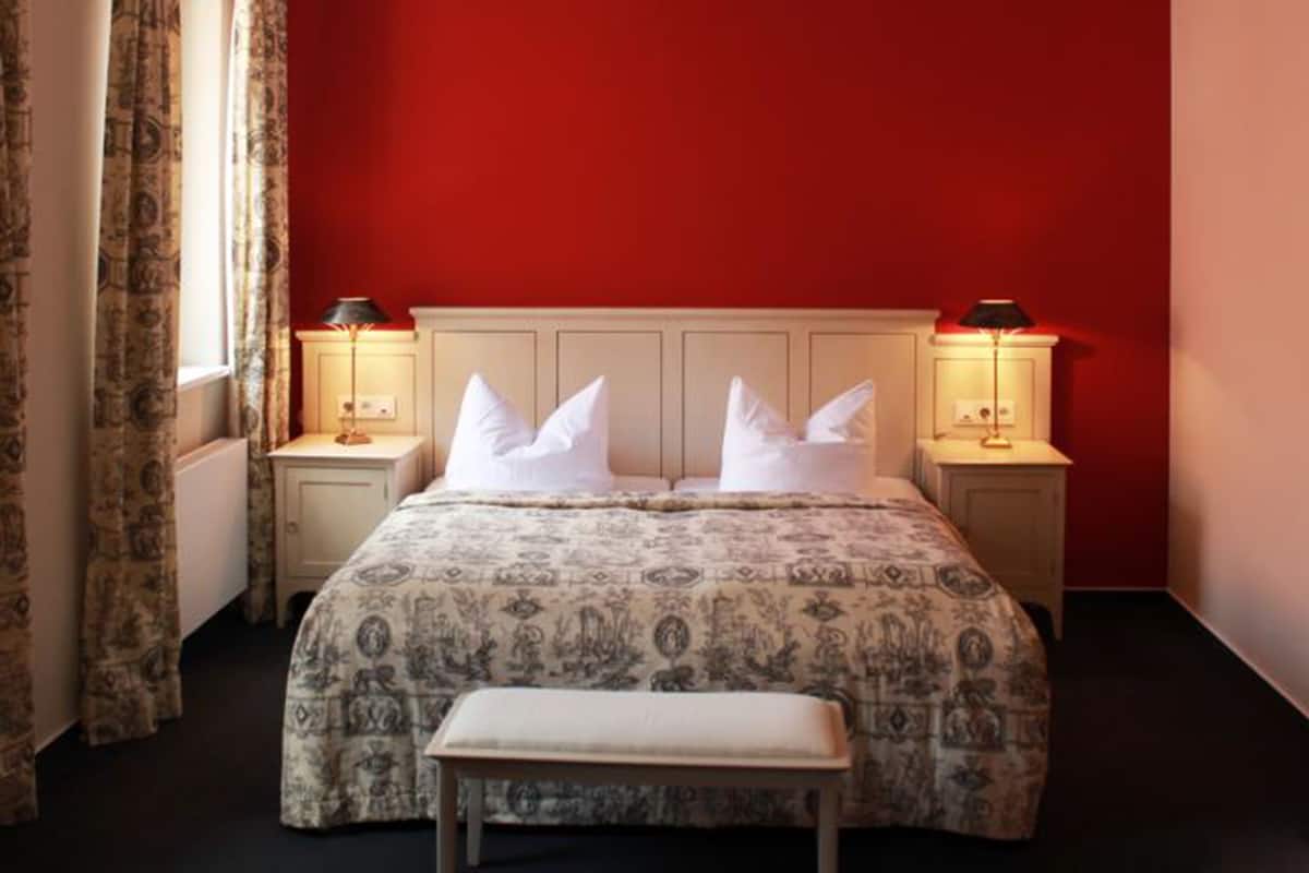Une chambre avec un mur rouge, une couvre-lit à motifs, deux lampes de chevet et un banc.