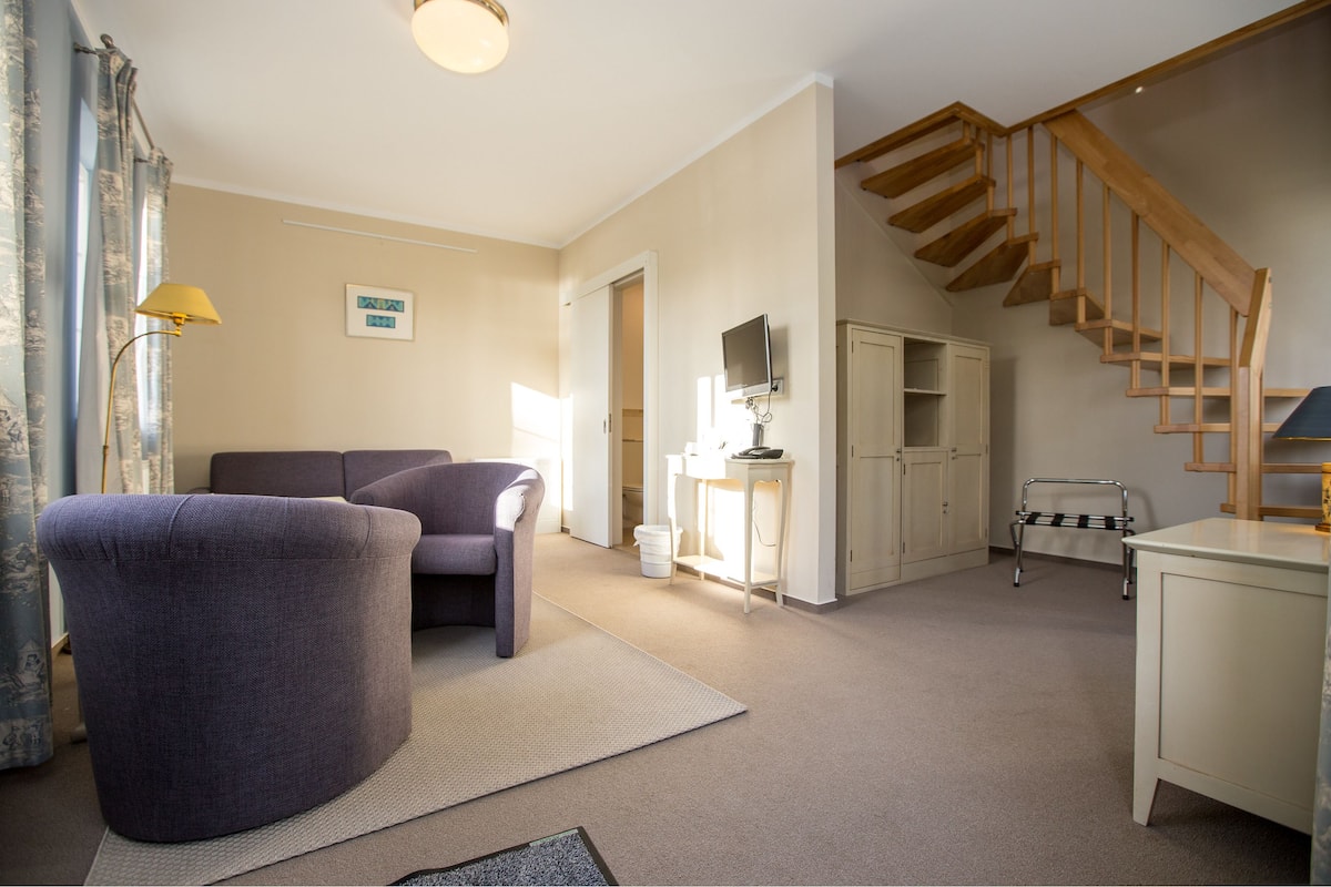 Un salon spacieux comprenant un canapé, un pouf rond, une lampe, un escalier et une petite table.