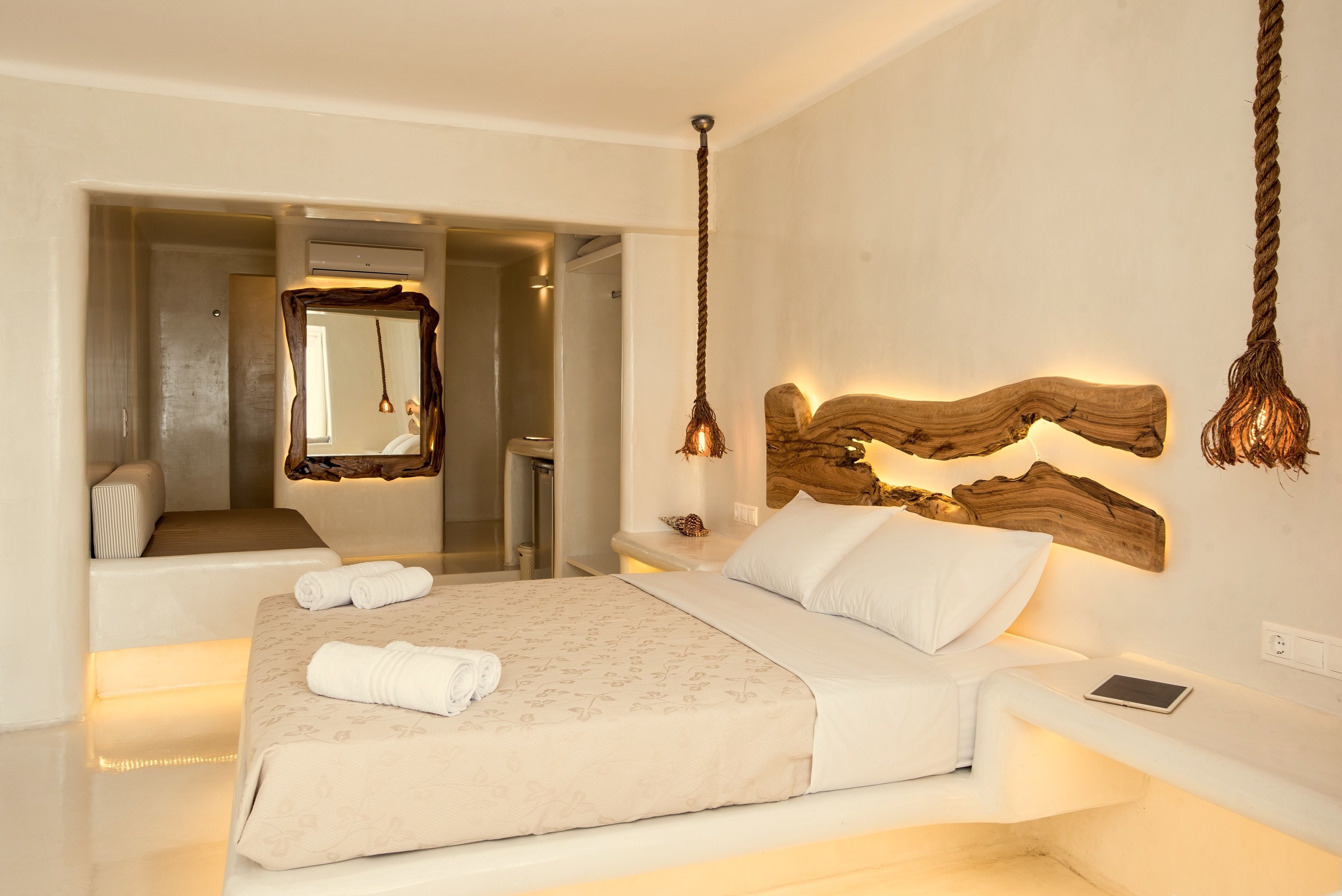 Una habitación de hotel moderna con una cama grande, una mesita de noche con una tableta, un espejo de marco original y un cabecero de madera con luces colgantes.
