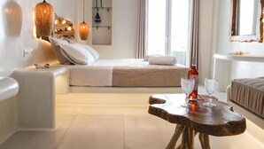 A hotel room with a bed, bedside table, and a wooden table with two glasses. - Villa Elina Suites And More (Mykonos)