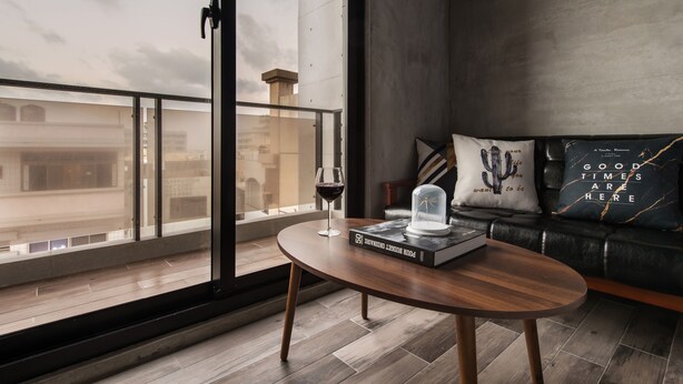 Una sala de estar moderna con un ventanal grande, una mesa de centro redonda de madera, un libro, una copa de vino y un sofá de cuero negro con cojines decorativos.