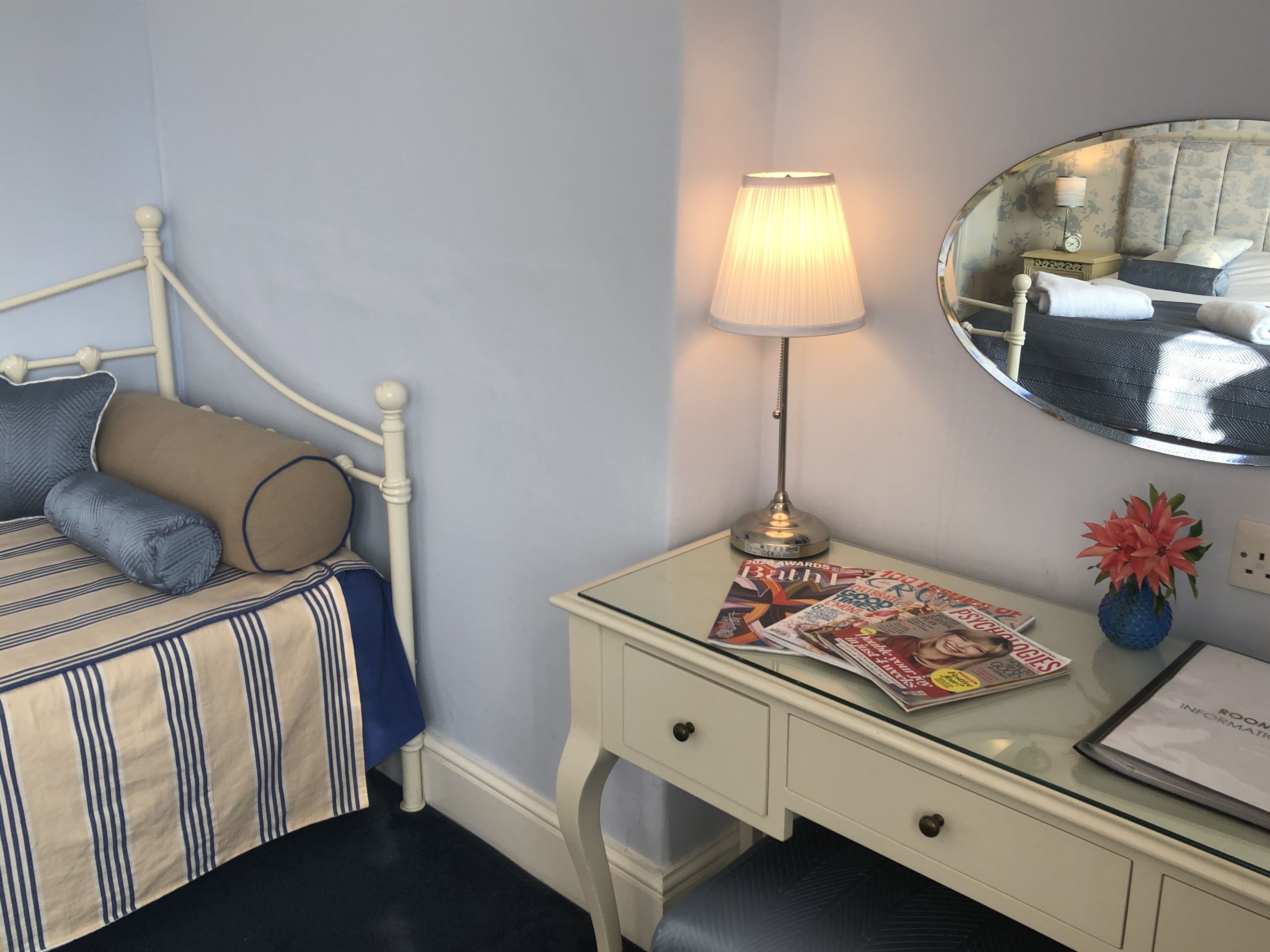 Un lit bien fait, recouvert d’une couverture à rayures, une lampe de chevet, un miroir et un bureau avec un magazine et un petit vase.