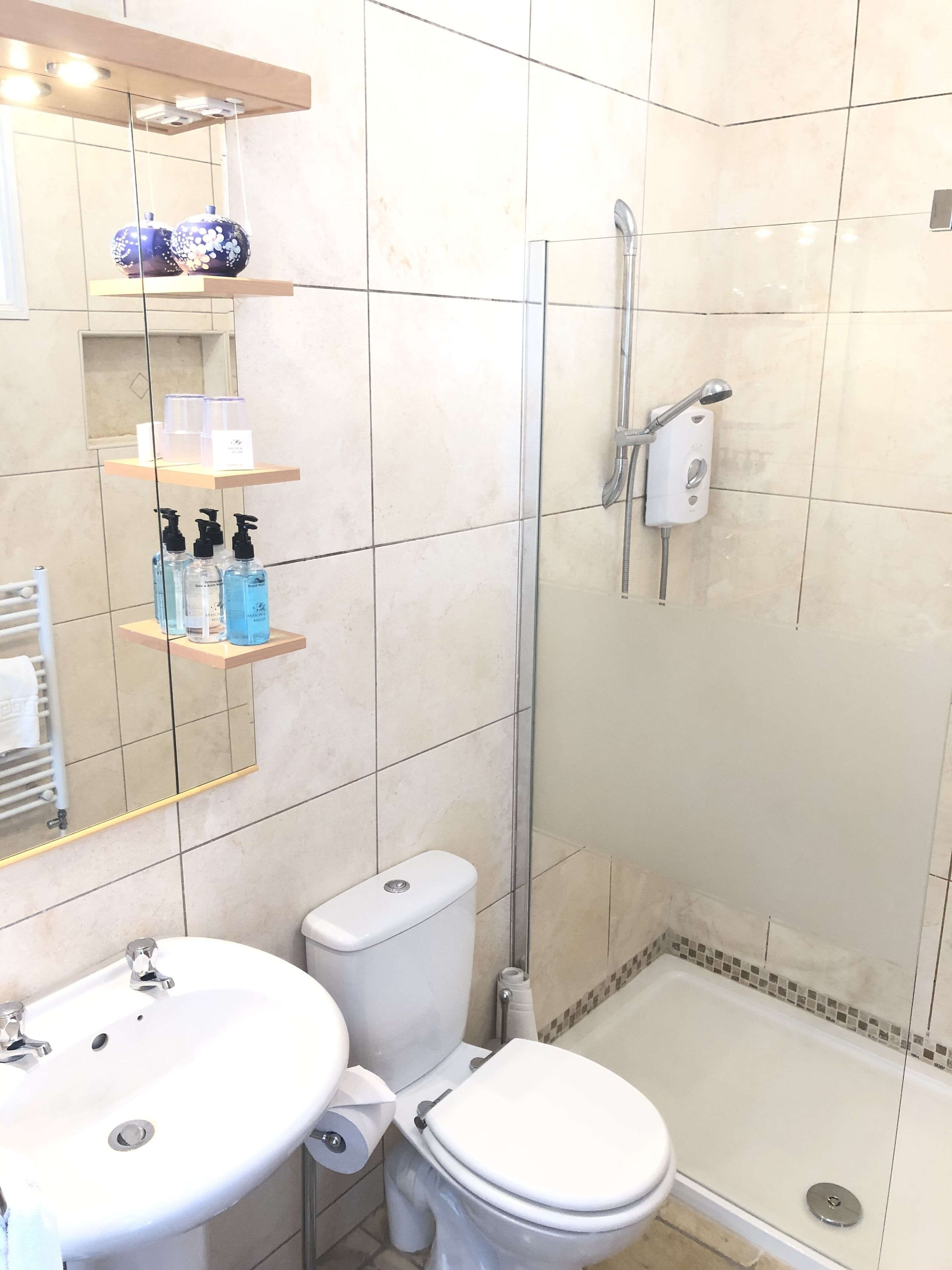 Ein Badezimmer mit Toilette, Waschbecken und Dusche. Regale mit Pflegeprodukten und ein Spiegel über dem Waschbecken.