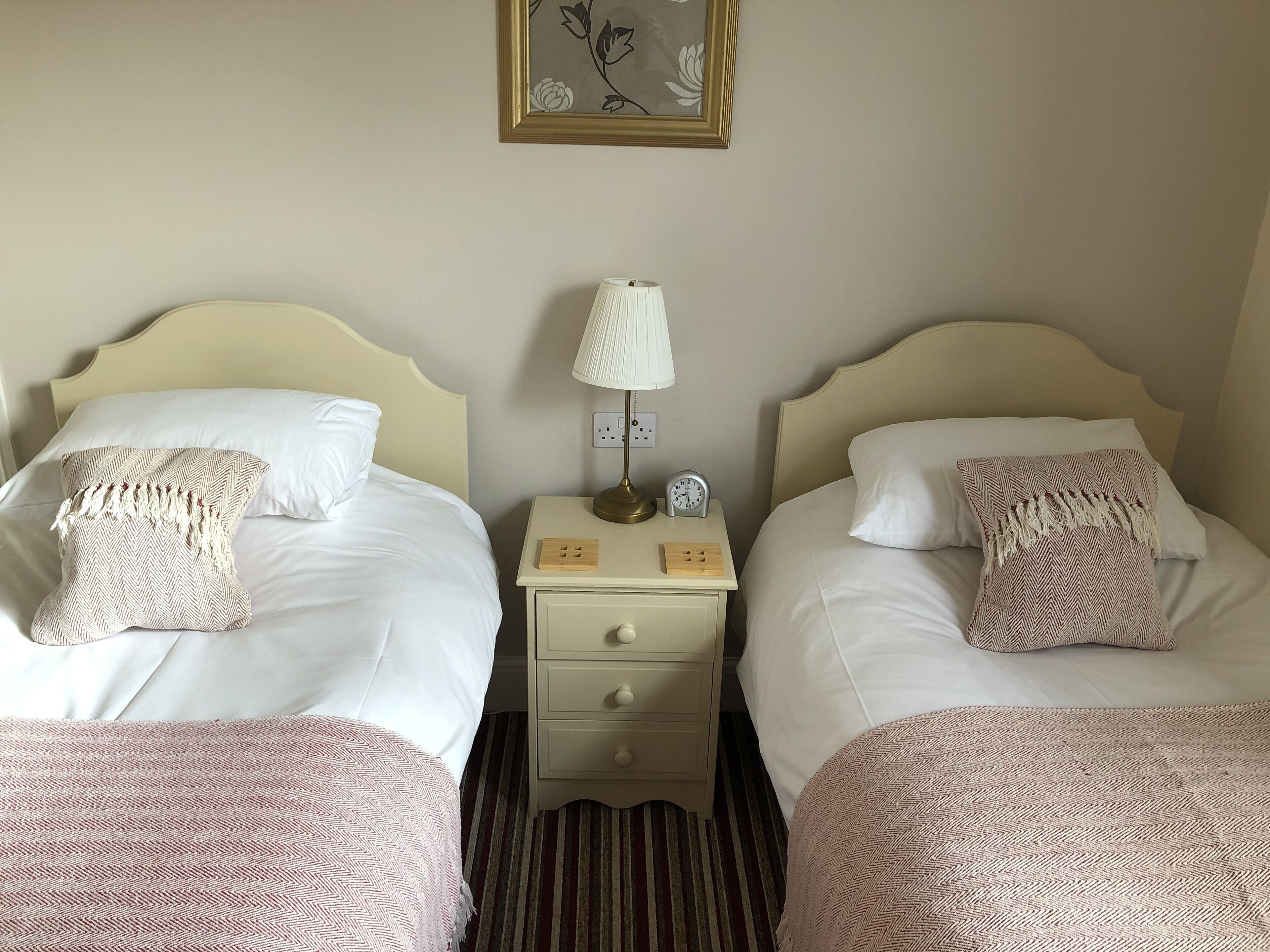 A hotel room with two beds, each with a white headboard, a bedside table with a lamp, and a small clock.