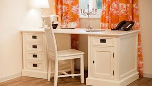 A white desk with drawers, a chair, a candelabra, a telephone, and a window with orange curtains. - Café Wildau - Pension am Werbellinkanal (Schorfheide)