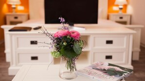 A room with a white bed, a TV on the wall, a bedside table with a vase of flowers, and a magazine. - Café Wildau - Pension am Werbellinkanal (Schorfheide)