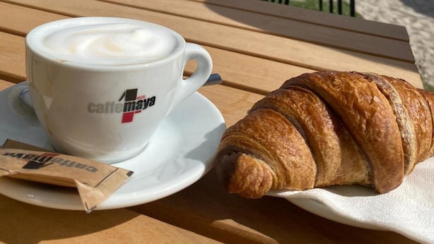 Un croissant et une tasse de café sur une table en bois, avec une vue sur la plage et l’océan en arrière-plan.