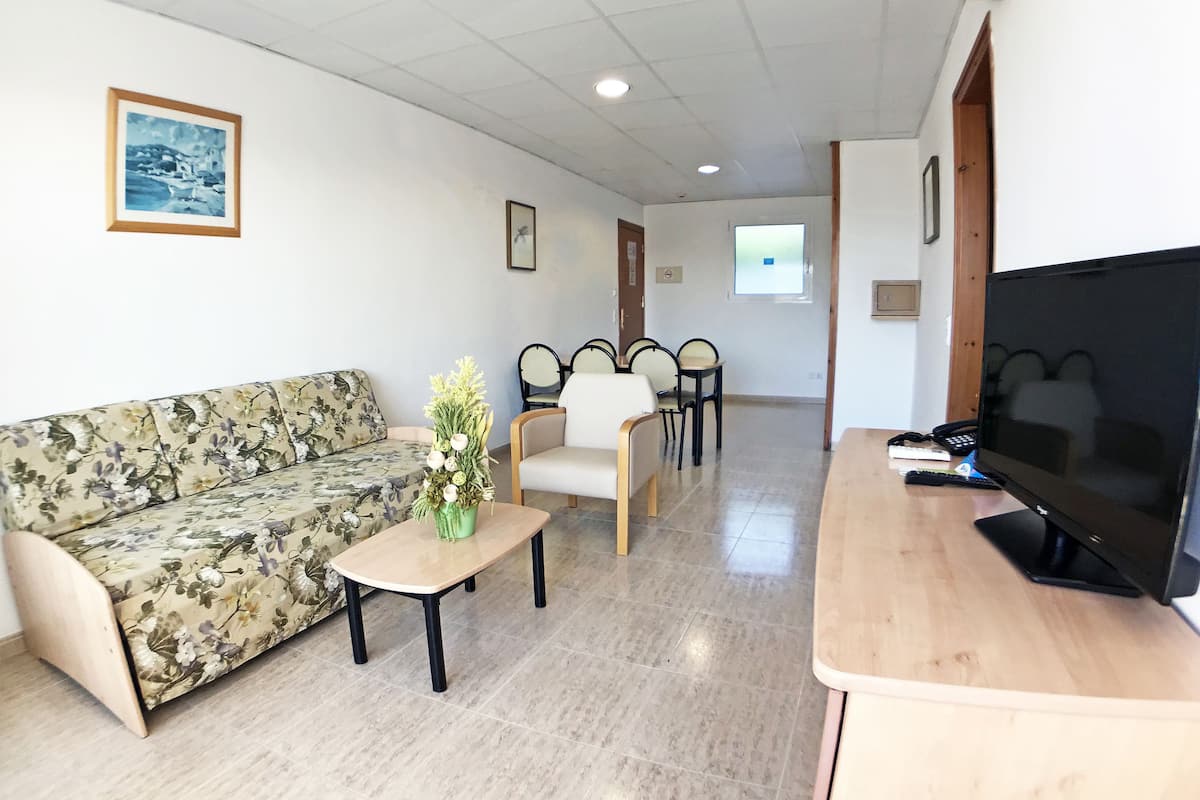 A hotel room with a floral sofa, a small table with a vase, a TV, and a seating area with chairs.
