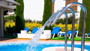 Una piscina con agua brotando de una fuente, rodeada de sillas de descanso y un edificio de fachada blanca.