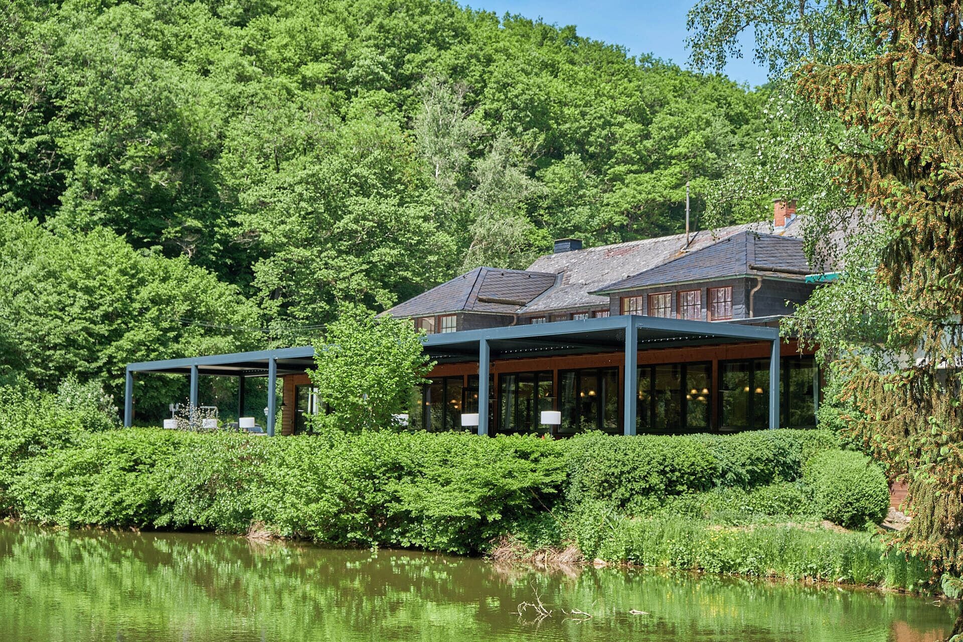 Un bâtiment situé au bord du lac, doté d’une façade en verre, entouré d’une végétation luxuriante et surplombant un lac paisible.