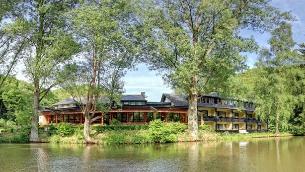 A lakeside hotel with multiple buildings, including a main lodge and guest cabins, surrounded by trees and a calm lake. - Hotel Forellenhof (Bundenbach)