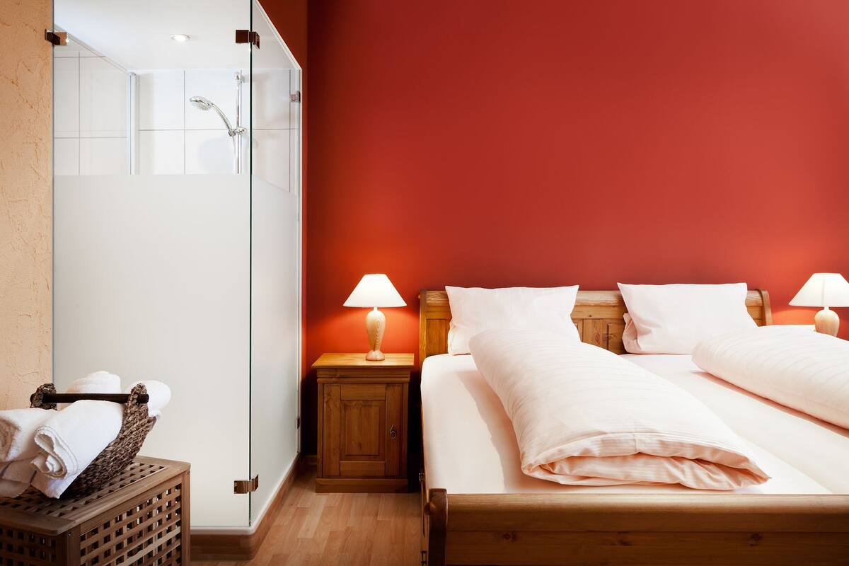 A hotel room with a wooden bed, two bedside lamps, a wooden nightstand, a glass shower enclosure, and a wicker basket with towels.