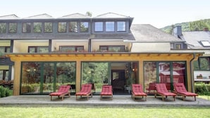 A modern building with a covered patio area featuring six red lounge chairs. - Hotel Forellenhof (Bundenbach)