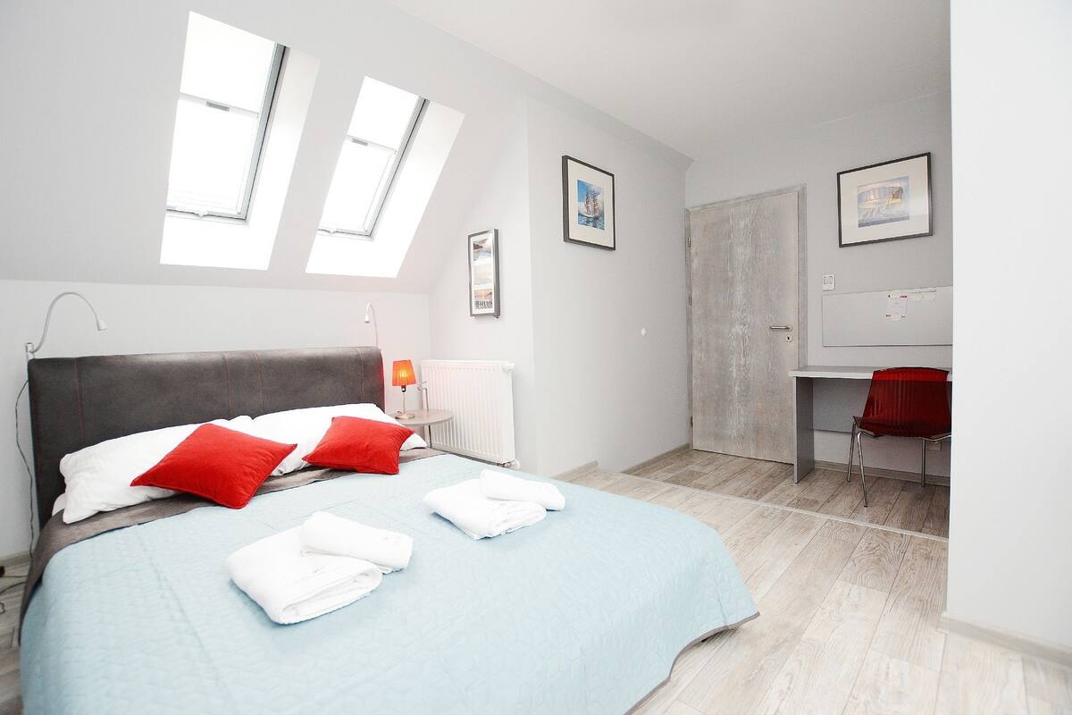 A modern bedroom with a bed, two windows, a desk, and a chair.