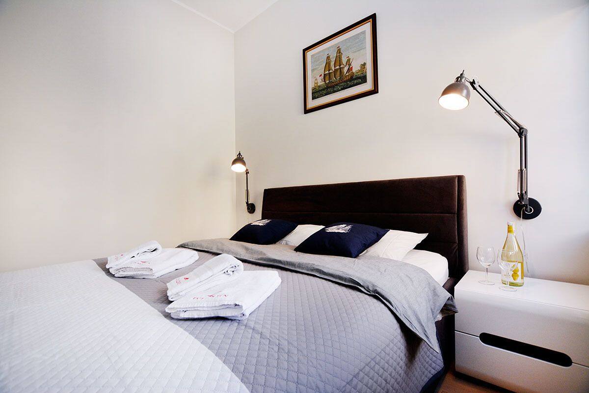 A neatly made bed with a grey quilt, two pillows, and folded towels on a white nightstand. A bottle of wine and a glass are on the nightstand.