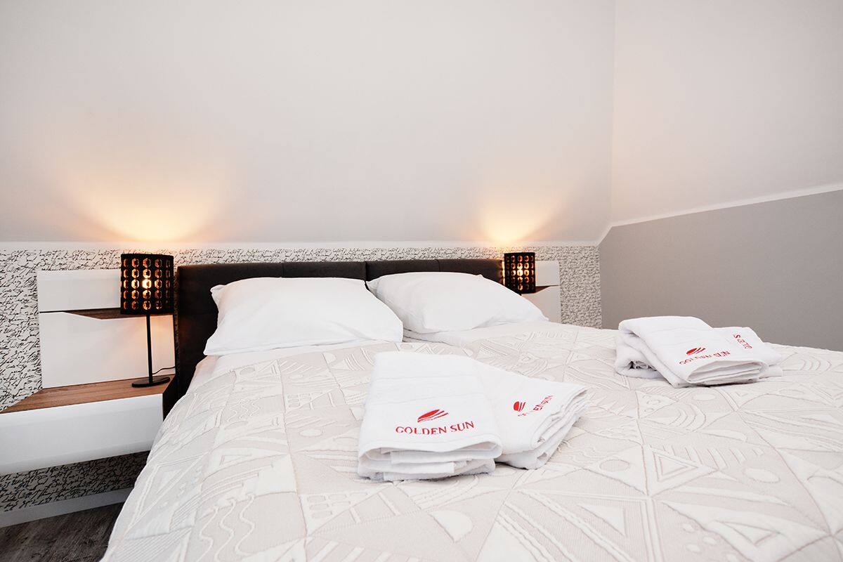 A neatly made bed with white linens and towels, a headboard with a textured design, and two bedside lamps with a patterned shade.