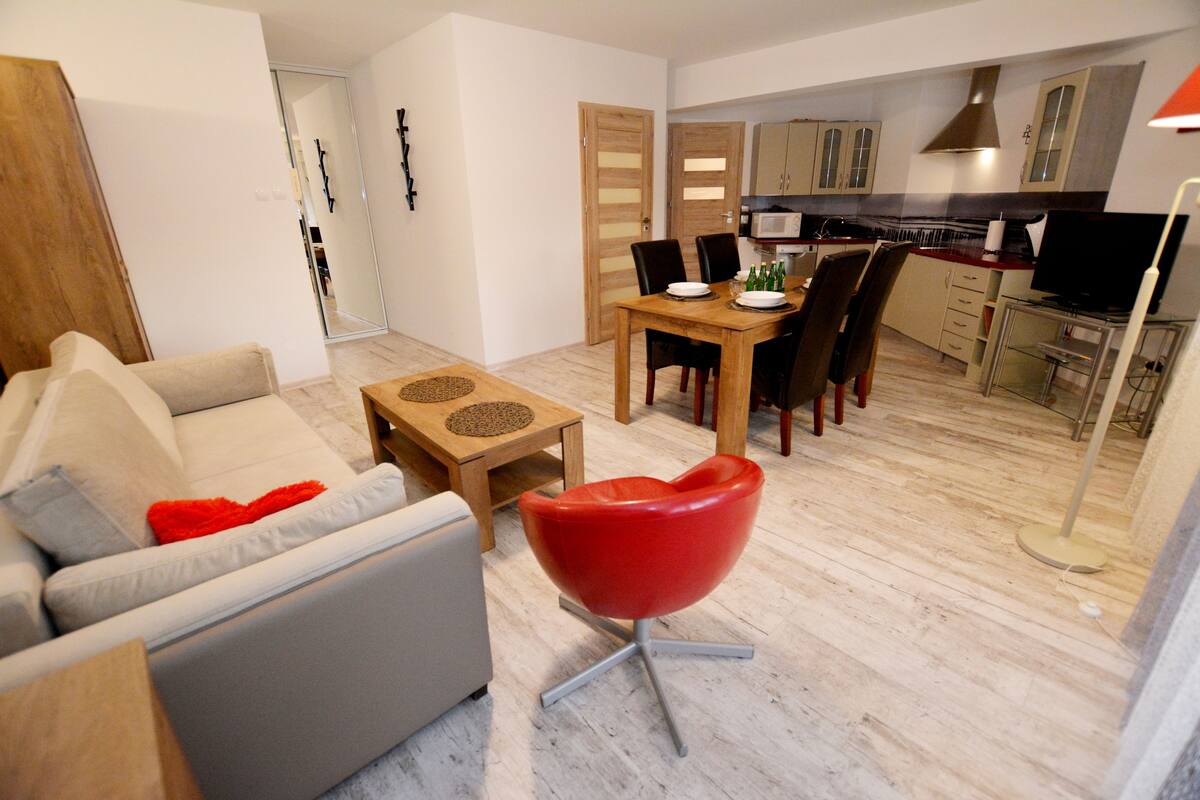 A modern living room with a sofa, a red chair, a dining table with chairs, and a kitchen area with cabinets and a microwave.