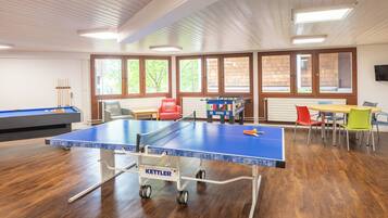 A ping pong table with a Kettler brand, a foosball table, and a pool table in a room with wooden floors and large windows.