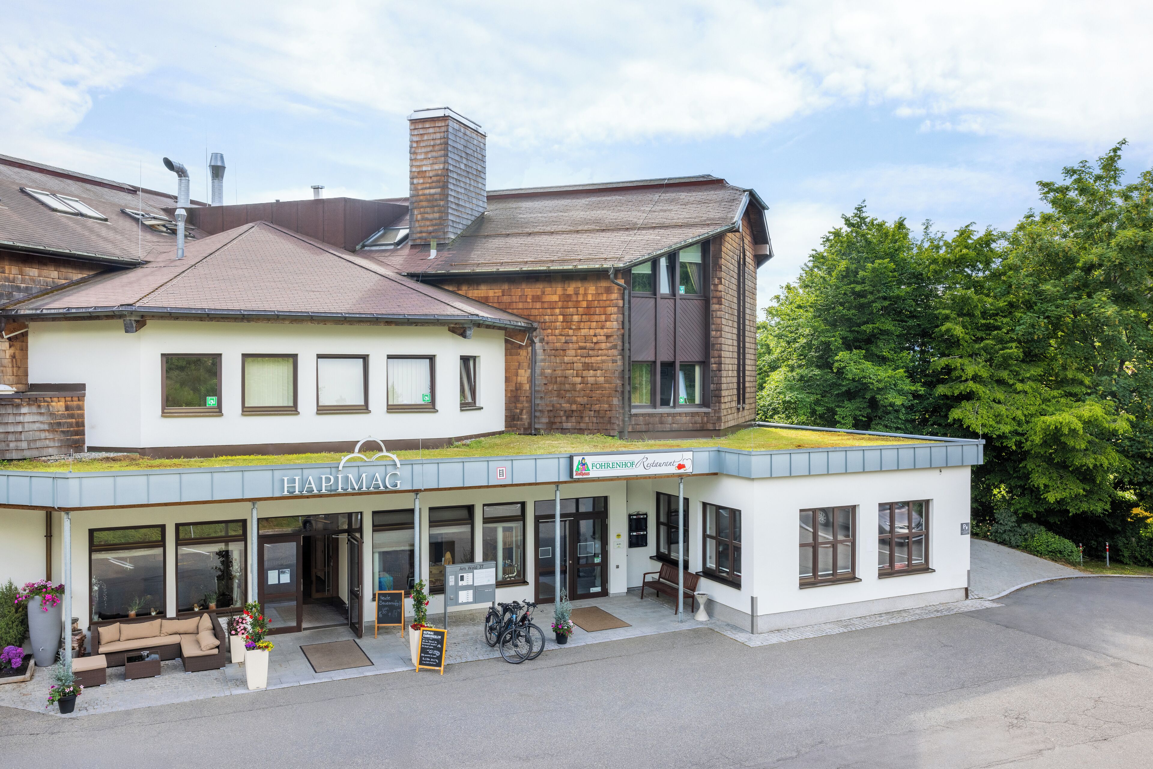 Un hotel con techo verde, zona de cafetería con mesas al aire libre y un letrero que dice "HAPIMAG".