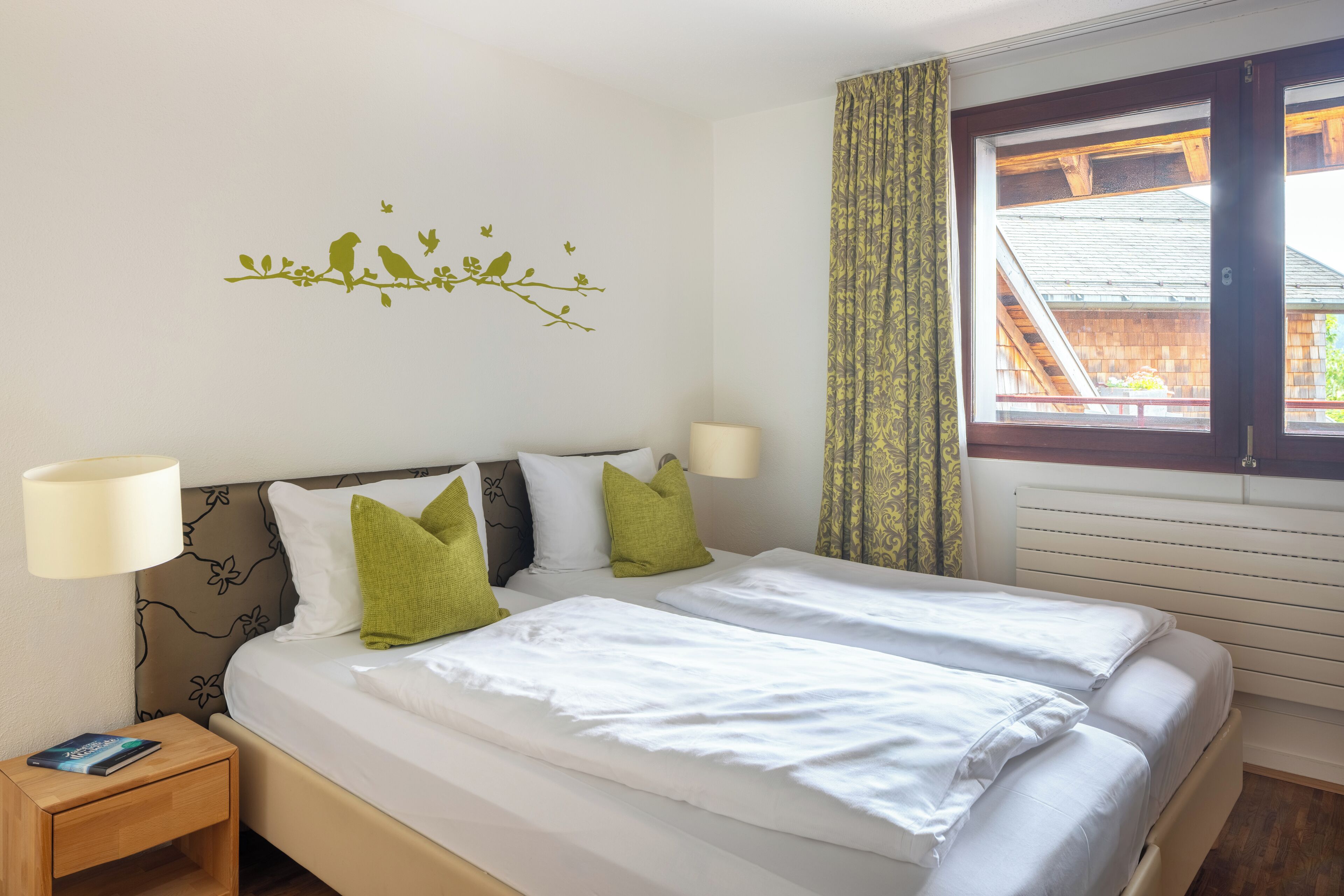 A double bed with white linens and green pillows, a nightstand with a lamp, and a wall decal of birds on a branch.