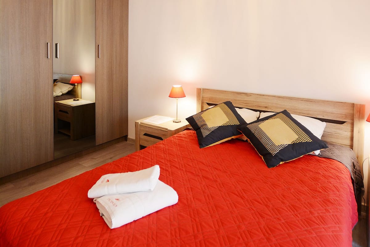 A bedroom with a large bed, a wooden wardrobe, a nightstand with a lamp, and a chandelier.