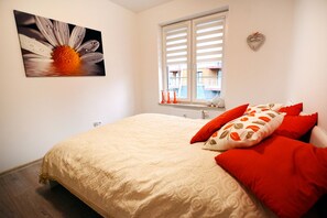 A bedroom with a large bed, a window with blinds, a wall-mounted picture, and a heart-shaped decoration.