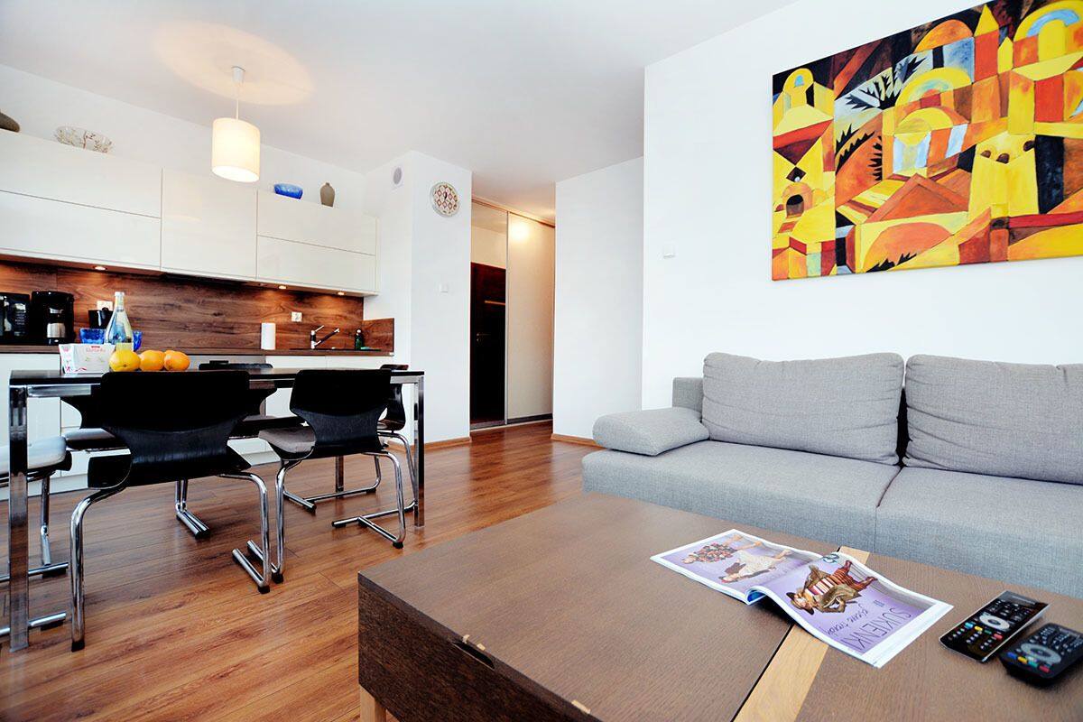 A modern living room with a grey sofa, a wooden coffee table with a magazine and remote controls, and a kitchen area with white cabinets and a wooden countertop.