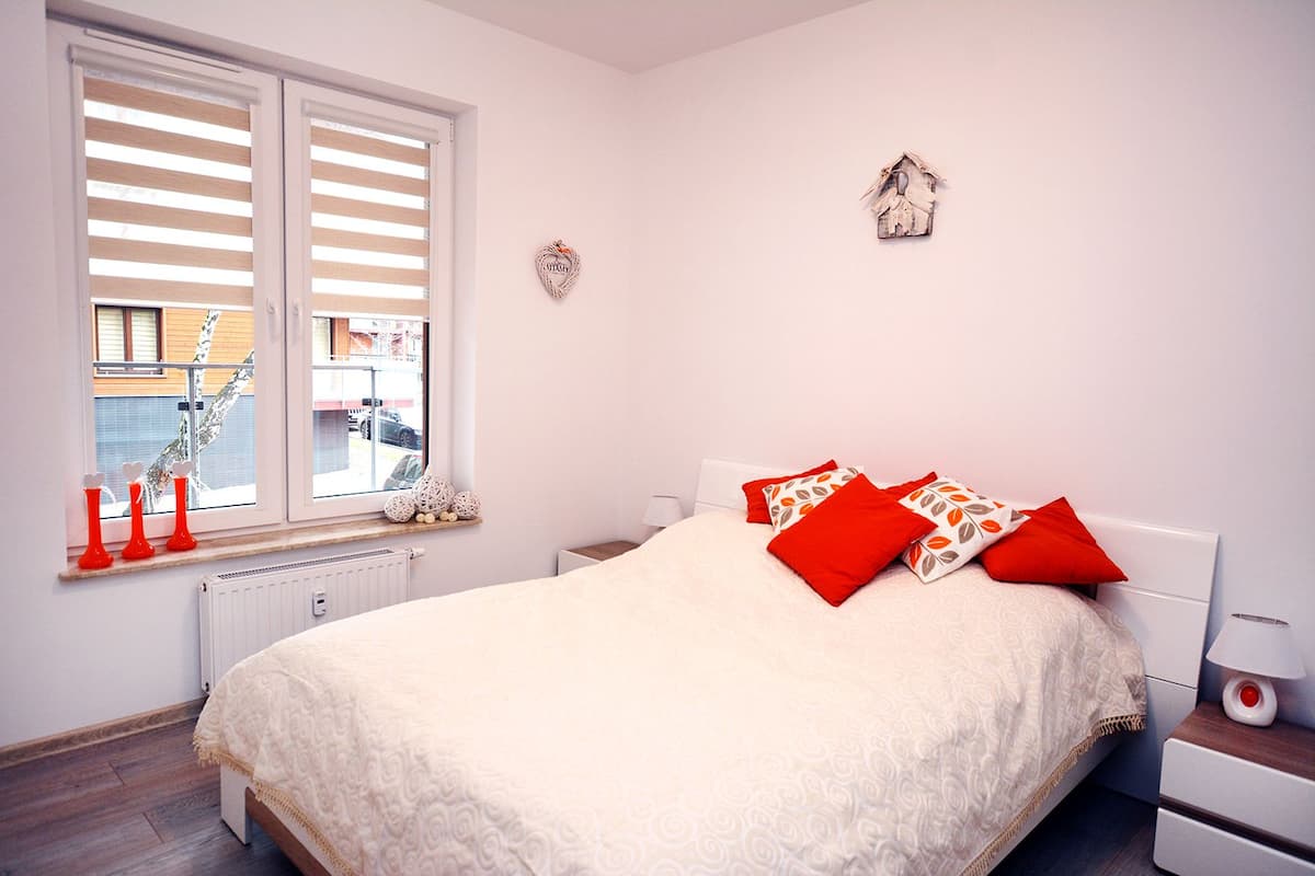 A bedroom with a large bed, a window with blinds, a bedside table with a lamp, and decorative items on the windowsill.